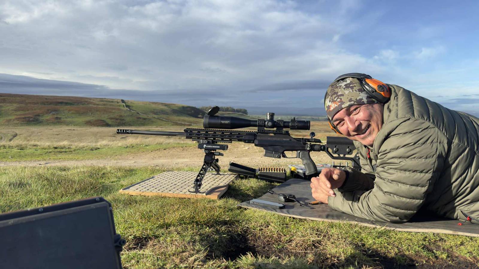 A precision rifle on a bipod at an open moorland range, with a chronograph case in the foreground and the shooter prone behind it.
